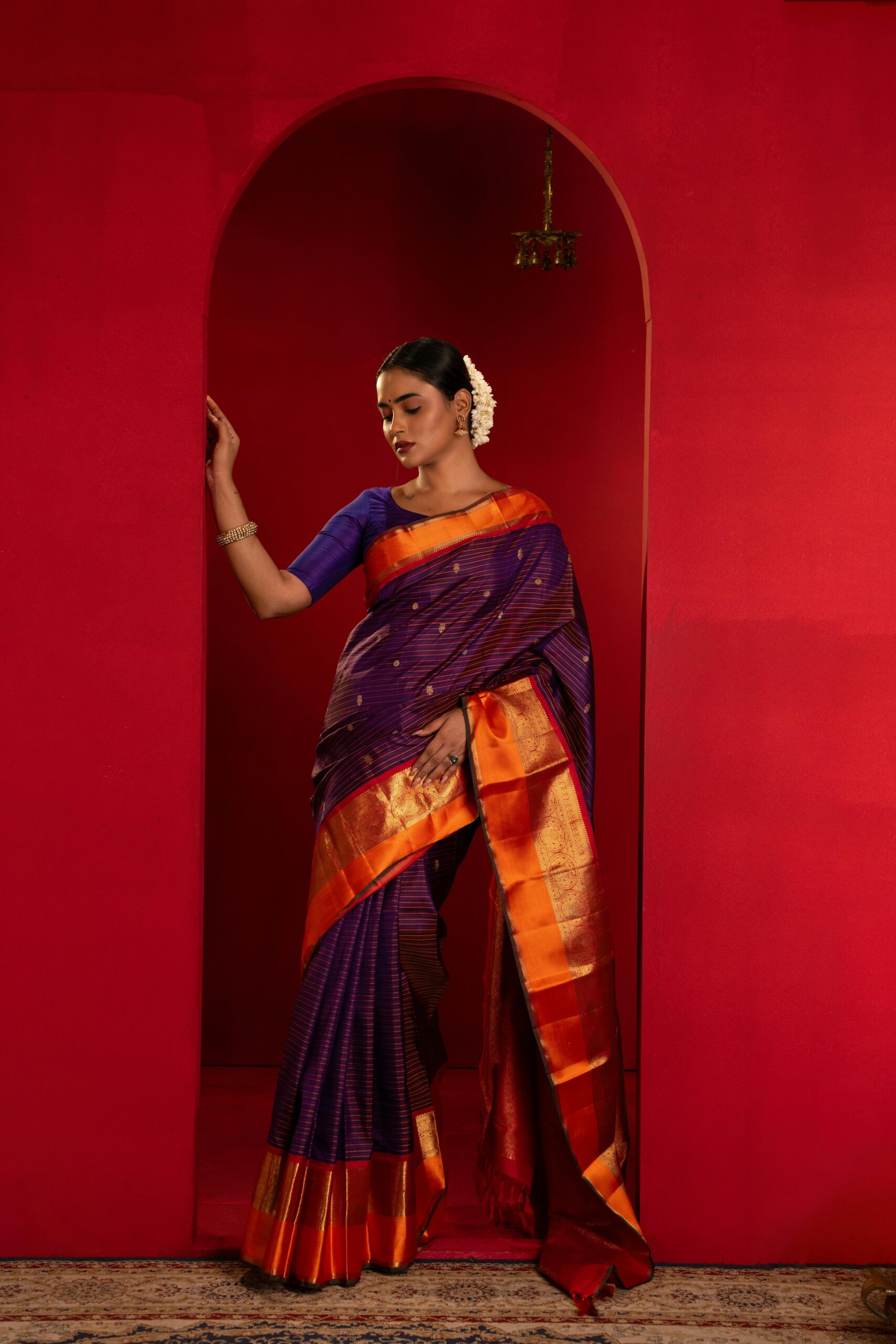 A woman gracefully poses in a vibrant Indian saree with a red background, showcasing traditional fashion.